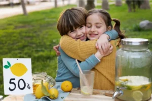 Duas crianças se abraçando em uma tenda de venda de limonada, simbolizando os primeiros passos dos empreendedores jovens.